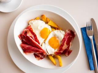Traditional appetizing spanish lunch - fried eggs with french fries and slices of jamon on white plate