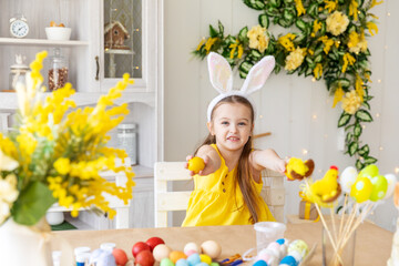 Obraz na płótnie Canvas Portrait of funny kid girl with plush bunny ears on head has fun with multicolored Easter eggs while sitting table in cozy kitchen at home,. Easter activities for kids concept