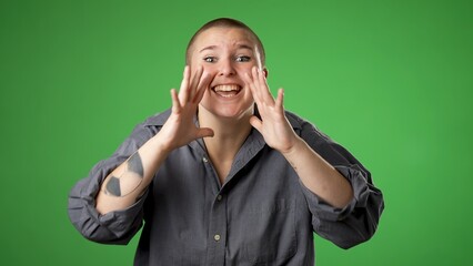 Portrait of happy gender fluid non binary young woman 20s smiling waving shout calling inviting with hands at mouth say hey you isolated on green screen background.