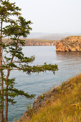 Warm summer foggy evening on shore of Baikal Lake at sunset. View from Olkhon Island to Small Sea. Natural background with larch trees on  rocky shore. Picturesque landscape. Summer travel and hiking