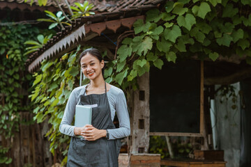 beautiful barista in apron holding the digital tablet while standing outside the coffee shop with the plants at the background