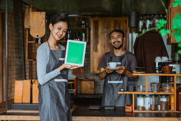 female barista in apron standing showing the digital tablet with green screen with the male barista standing at the background holding a cup of coffee