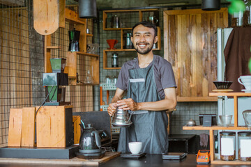male barista in apron standing with smile while brewing the coffee inside the bar desk