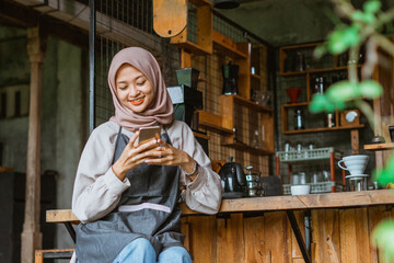 female barista with hijab and apron using the phone while sitting in front of the bar desk