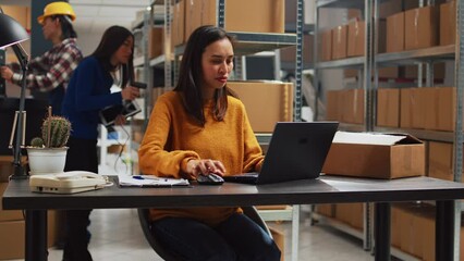 Young person doing stock inventory with laptop, writing logistics information on papers for business development. Female employee planning products distribution in warehouse space. - Powered by Adobe