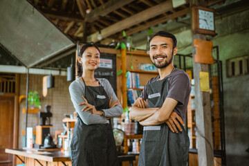 a couple of barista in apron standing together with handcrossed and smiling inside the coffee shop