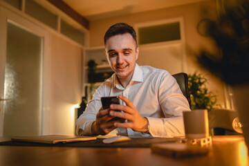 A young man is working late or studying at his home office while drinking coffee and using his mobile phone	