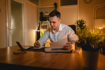 A young man is working late or studying at his home office while drinking coffee and using his mobile phone	