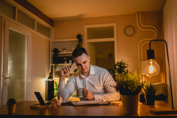 A young man is working late or studying at his home office while drinking coffee and using his mobile phone	