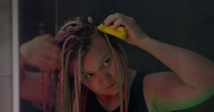 Young woman with multicolored box braids hairstyle is trying to wash her scalp, using a sponge and shampoo, while standing in the shower in the semi-darkness, front view.