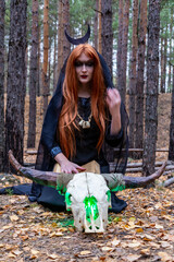 a girl in a Gothic image for Halloween is sitting in the forest with a cow's skull in the autumn forest in the background is a pine forest