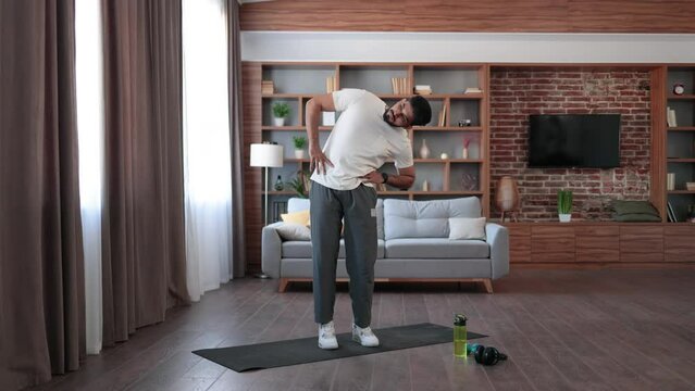 Full length portrait of active indian man doing side tilts with hands on hips at cozy living room. Bearded young guy in sport clothes having regular domestic workout.