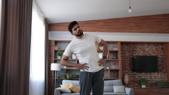 Arabian bearded man dressed in sport clothes holding hands on hips and bending body to sides. Sporty young athlete spending free time for domestic workout.