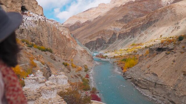 Beautiful landscape of Phugtal monastery in remote Himalayan mountains with flowing Tsarap chu river. Rack focus to the tourist girl wearing hat and enjoying view of nature. Girl in the mountains. 