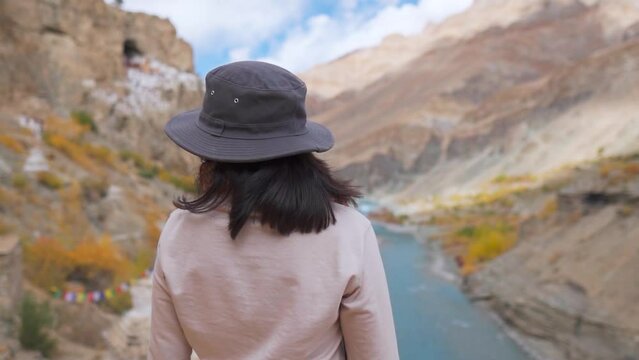 Young Indian woman standing against Phuktal, Phugtal Monastery and Tsarap Chu river at Zanskar, India. Rack focus to the nature background from tourist female wearing hat. Travel and holiday concept. 