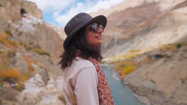 Portrait of beautiful Indian girl in hat standing against Phugtal monastery and Tsarp Chu river in mountains of Zanskar, Ladakh, India. Tourist enjoying view of beautiful nature during holidays.