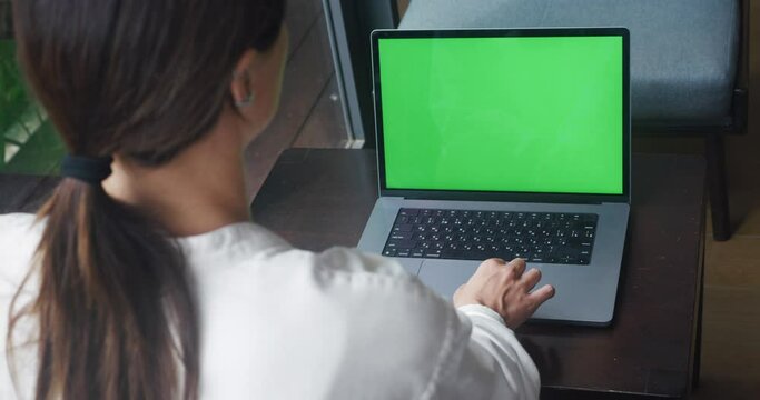 Freelancer female designer uses laptop with green screen mockup in cozy cafe in front of window. View from behind the shoulder female uses a computer with green screen chroma key in stylish interior