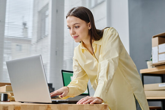 Young female volunteer bending over workplace with laptop while looking at screen and scrolling through onlinr requests of people in crisis
