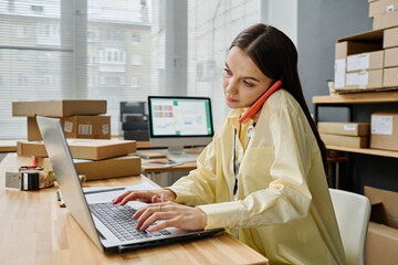 Busy young brunette female volunteer sitting by workplace in front of laptop and talking on mobile...