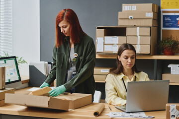 Two young female volunteers taking online requests of people in need and packing boxes with...