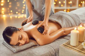 Woman getting exotic spa massage with hot stones. Happy, relaxed young woman lying on spa bed while professional masseuse is putting hot stones on her back. Spa treatment, body relaxation concept