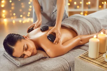 Papier Peint photo Spa Woman getting exotic spa massage with hot stones. Happy, relaxed young woman lying on spa bed while professional masseuse is putting hot stones on her back. Spa treatment, body relaxation concept