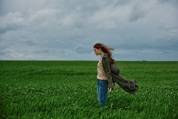 Fototapeta premium a sad woman in a coat stands in a field in cloudy weather resisting a strong wind