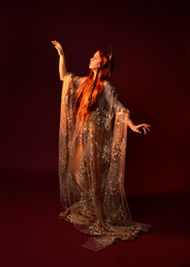 Full length fantasy portrait of beautiful woman model with red hair, goddess silk robes & gold crown. Standing pose gestural hands reaching out isolated on dark red studio background 