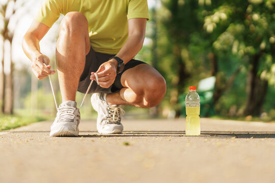 Young athlete man tying running shoes with Energy Drink water, male runner ready for jogging outside, asian Fitness walking and exercise in the park morning. wellness, wellbeing and sport concepts