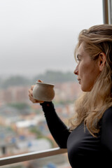 beautiful blonde woman having a coffee on a rainy and cloudy afternoon seen from the window with a cup of coffee in her hand happy and thoughtful