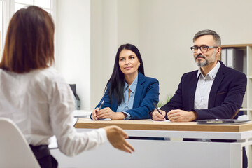 Fototapeta premium Young brunette woman at interview and two business partners listening attentively to their potentially new employee. Three people at meeting in bright modern office. Candidate for position in company.