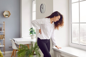 Woman suffers from lumbago pain. Young girl standing by the window sill at home, feeling pain, and holding her hand on her lower back. Backache, health problems, concept - obrazy, fototapety, plakaty