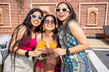 Three beautiful young indian girls wearing sunglasses having fun together holding glasses of drinks...
