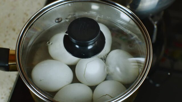 Pot of boiling water in which white chicken eggs are boiled. Water boils and boils in pan with boiled eggs. Female hand closes pan with boiled white chicken eggs with transparent glass roof. Close-up
