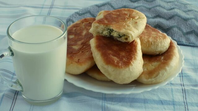On table, covered with gray tablecloth, there is glass of fresh milk and plate with ruddy homemade pies, one pie is broken in half and one can see the filling of cottage cheese with herbs. Close-up