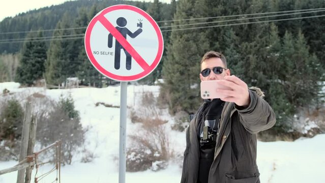 A man in sunglasses takes a selfie on the phone in front of the no selfie sign. A man takes a picture with his phone In a dangerous place