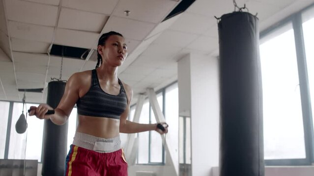 Young asian female boxer with braided hair fast and focused jumping rope next to punching bag warming up before boxing training in the indoor gym