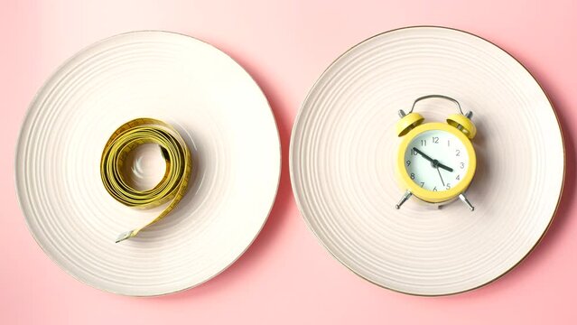 Female hands with an alarm clock and an empty plate with a centimeter tape on a pink background close-up top view.