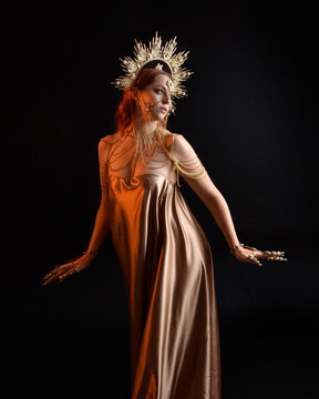 Close up fantasy portrait of beautiful woman model with red hair, goddess silk robes & ornate gold crown.  Posing with gestural hands reaching out, isolated on dark  studio background 