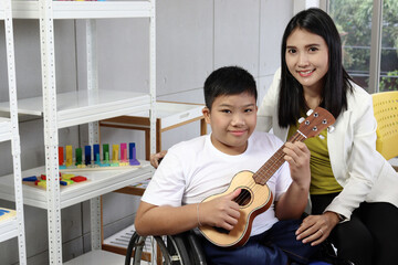 Disabled kids classroom, happy schoolboy on wheelchair learning to play ukulele with teacher, student have fun together during study at school, kid education with physical disability and intellectual.