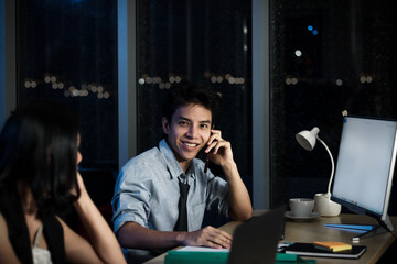 Happy smiling young Asian officer talking with mobile phones at working desk with colleague at night, staff business man  work hard overtime at night, overwork project overnight, hard-working worker