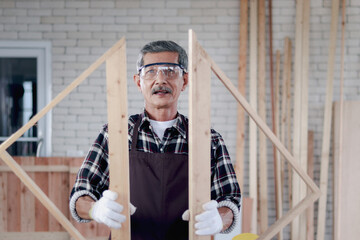 Portrait of senior Asian carpenter wear safety glasses and cloth gloves, holding a gable in woodwork carpentry, old citizen craftsman work in joinery workshop, elderly grandpa enjoy DIY hobby.