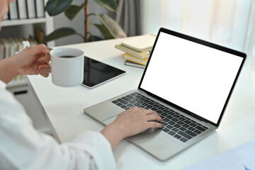 View from behind the shoulder young businesswoman holding coffee cup and working with laptop...