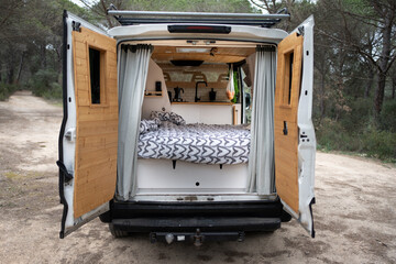 Back of camper with open doors, interior with wooden bed and kitchen, wooden doors, heart quilt and grey curtains, large white camper van, lonely forest interior. © Cristina