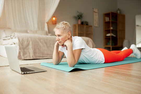 Side view image of good-looking mature female with short hair and slim body in sports clothes lying on floor on blue mat in her bedroom in front of laptop, watching attentively online fitness tutorial
