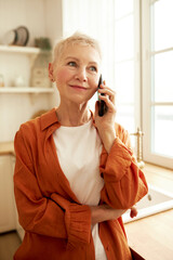 Vertical portrait of aged female in stylish orange shirt talking on phone to food delivery service, making order, standing against window at kitchen, looking aside with smiling face