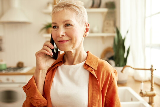 Happy mature lady with stylish short haircut having phone conversation standing at kitchen, talking to her beauty master, making an appointment for next week, listening attentively with smiling face