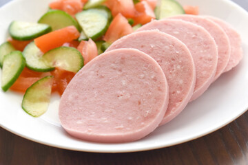 Greek Parizaki pork sausage slices with cucumber and tomato salad on a white plate. Very popular charcuterie products in Greece.  Sliced hams on a plate. 