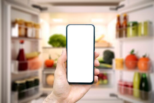 Online grocery delivery app in a mobile phone. The men is holding a phone in his hand opposite the refrigerator.Food market service in smartphone. Grocery shopping background concept.