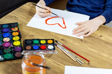 child creating drawing with multicolored paints and brush on wooden table close-up. Boy is payinting heart on white paper with watercolor